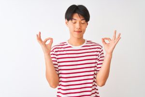 Young Chinese man wearing casual striped t-shirt, standing in front of plain white background, relaxing and smiling with eyes closed, doing meditation gesture with fingers closed in a circle. Image credit: AaronAmat (iStock). Image used in Faceyourfears.ca "How to Develop Self-Awareness (part 2): Meditation."