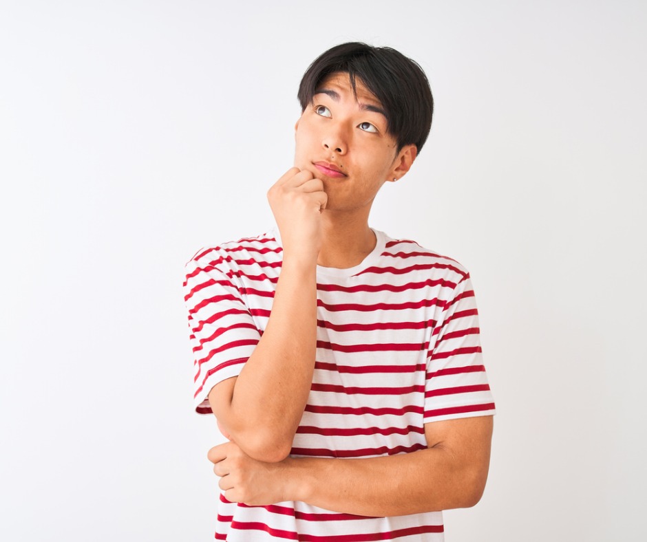 Young Chinese man wearing casual red striped t-shirt, standing in front of plain white background, looking thoughtfully upwards to his right, with one hand on his chin and other arm across his lower chest. Image credit: AaronAmat (iStock). Image used for faceyourfears.ca blog post “How to Develop Self-Awareness (Part 2): Meditation.”