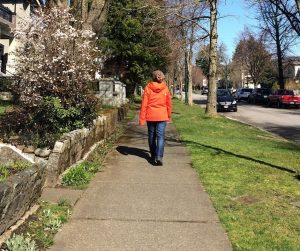 A woman (Mona Benjamintz) wearing orange jacket, jeans and a toque walking away from the viewer. Mona is walking on a sidewalk in a residential neighbourhood past houses, trees and under a blue sky on a sunny day. Image credit: Mona Benjamintz.
