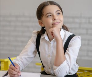 Young school girl with long brown hair worn in pig tails, wearing white blouse and dark jumper sitting at school desk, with pen in hand a notebook on the desk. She’s looking out the window, smiling and looking pensive. Her chin rests on her hand. Image by Zinkevych (iStock).