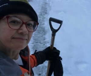 Smiling woman, looking at the camera, in orange jacket, mitts, tuque is standing outside on sidewalk partially cleared of show holding a shovel. The woman is the author of the blog post and is taking a break from shoveling show.